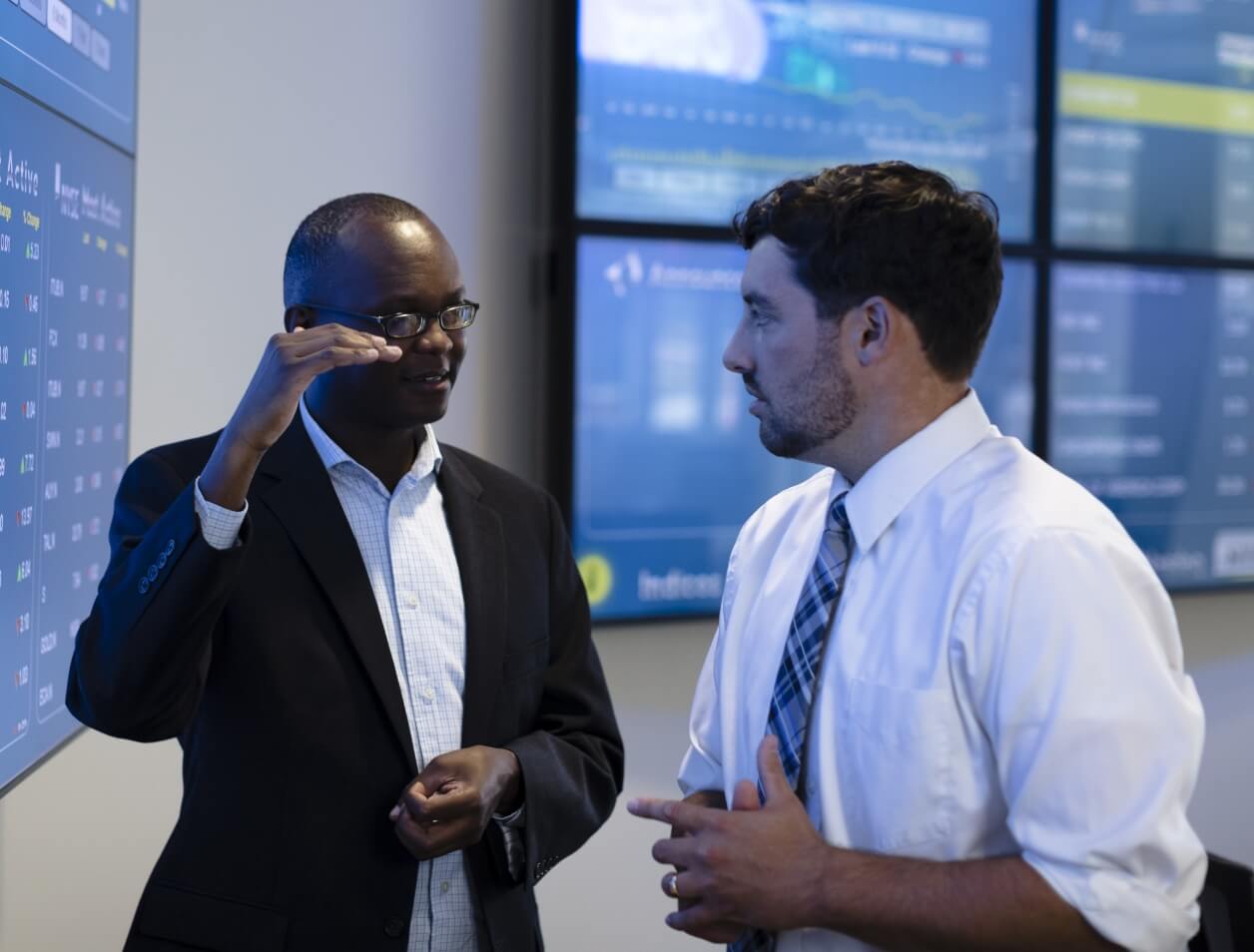 Two professionals discussing graphs displayed on monitors in a conference room